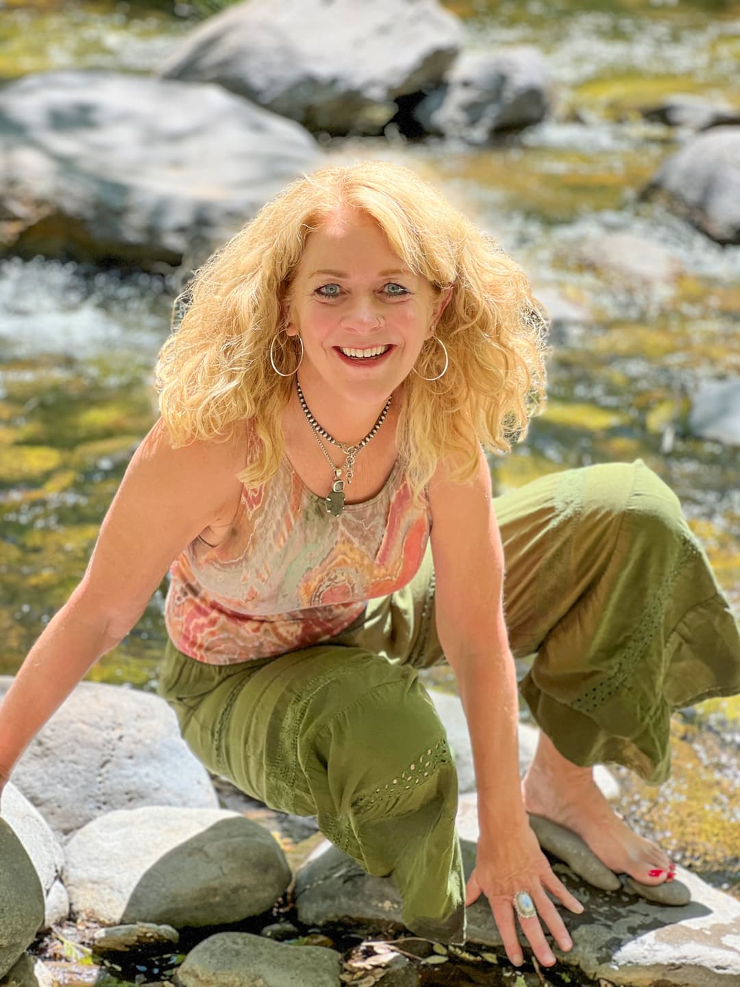 Amia Geismann, a Sedona-based transformational healer and spiritual guide, is pictured crouching peacefully on a rock in a flowing creek.